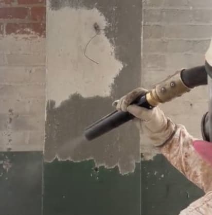 Operador dirige la boquilla de limpieza abrasiva contra un muro de concreto para retirar el recubrimiento viejo