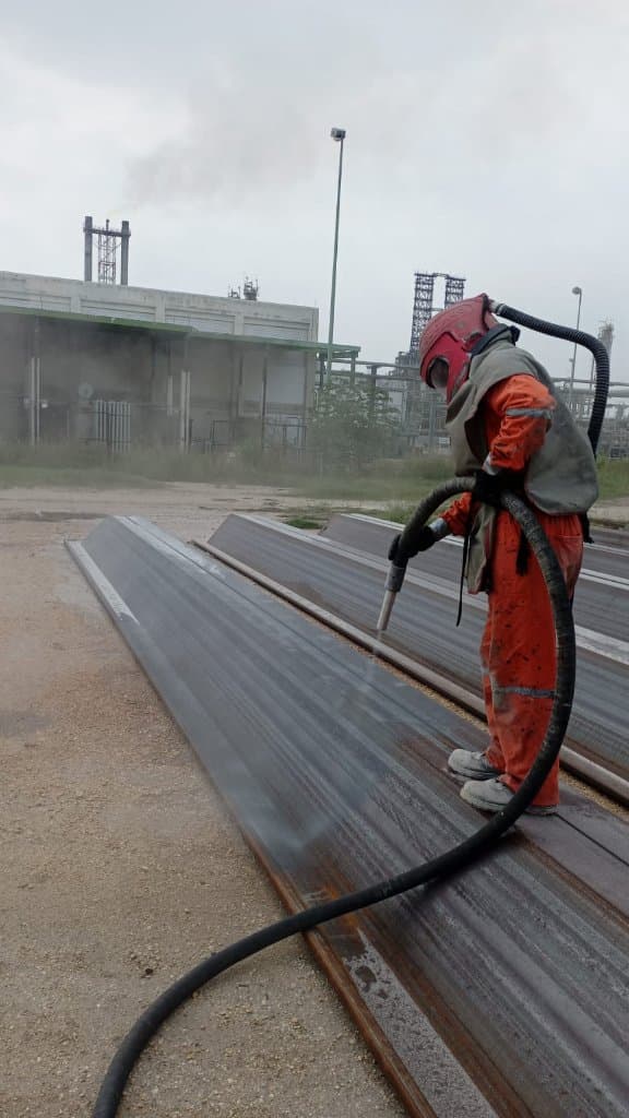 Operador con capucha de chorro limpia perfiles de acero oxidados en el patio de una planta petroquímica