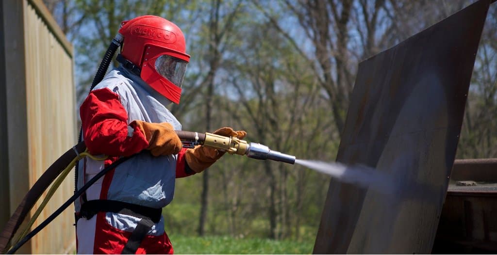 Operador con traje de protección y capucha de aire aplica chorro abrasivo sobre una placa de acero